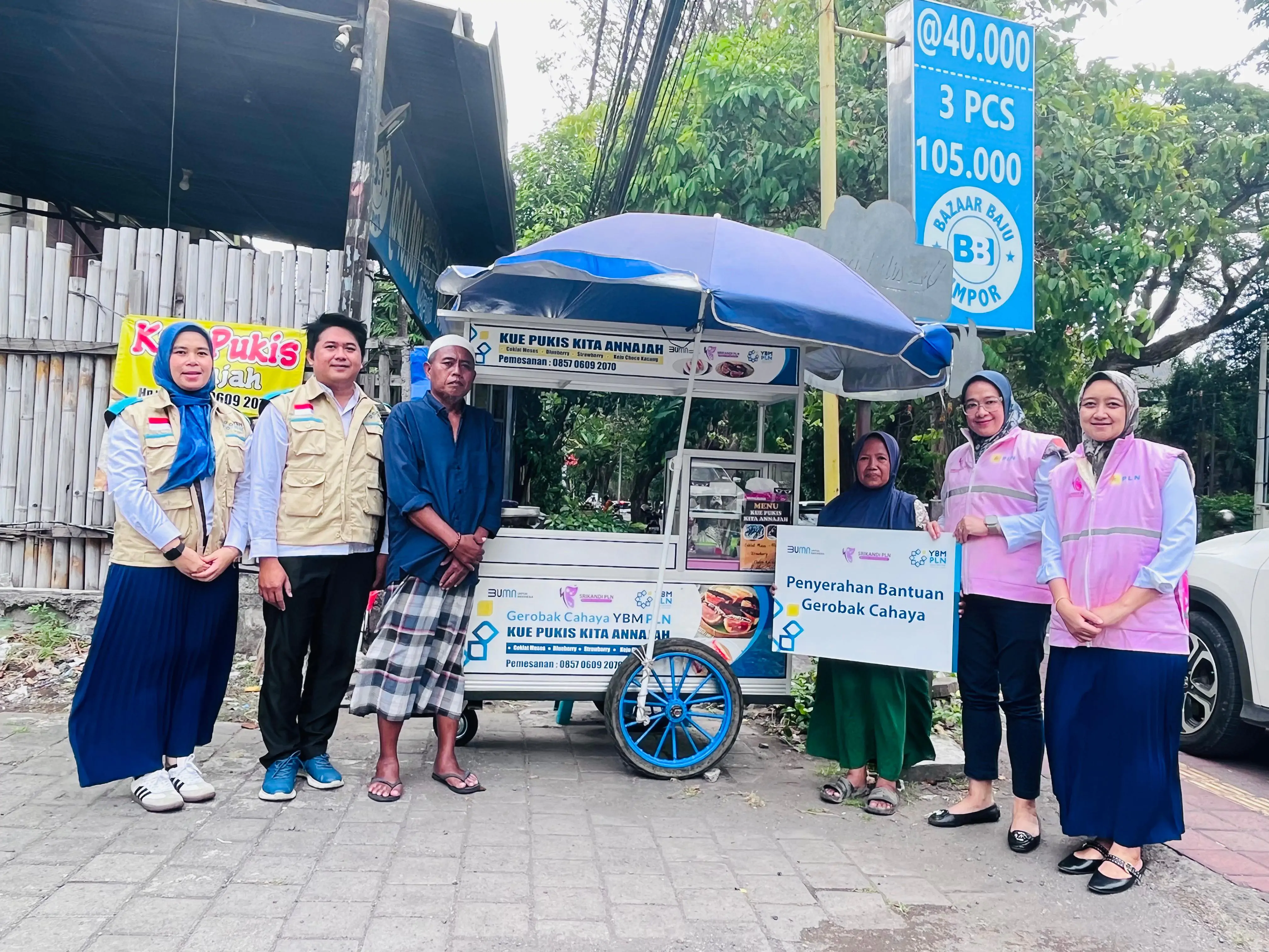 Penyerahan bantuan gerobak cahaya kepada Ibu Hayati pedagang kue pukis. Tampak Ibu Hayati memegang plakat simbolis Bantuan gerobak diapit Srikandi (rompi pink) dan Anggota YBM PLN UID Bali (rompi coklat) berpose didepan Gerobak Barunya.